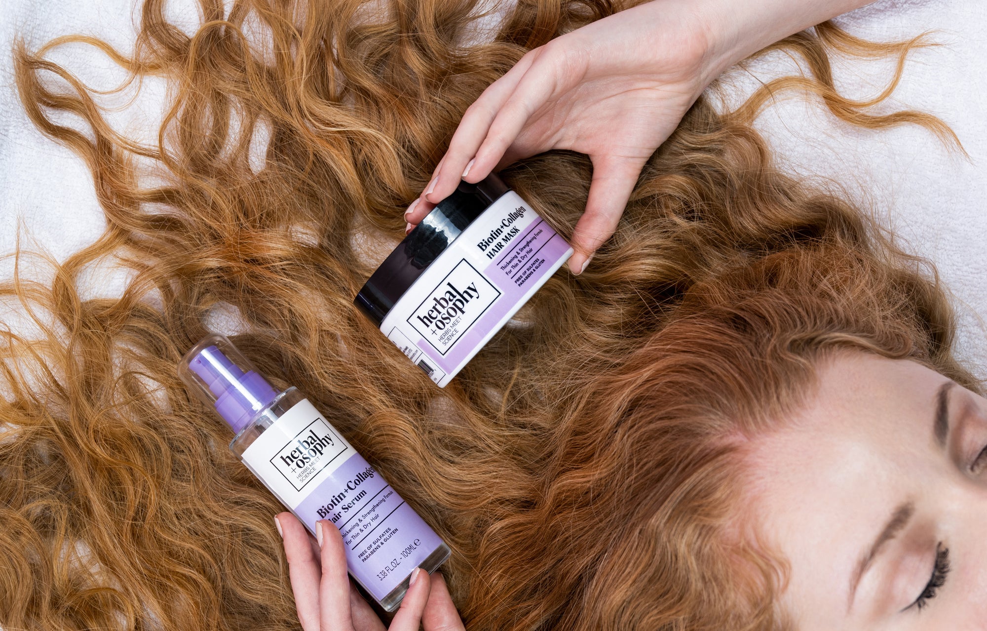 woman with long wavy hair holding Herbalosophy Biotin + Collagen serum and mask haircare products
