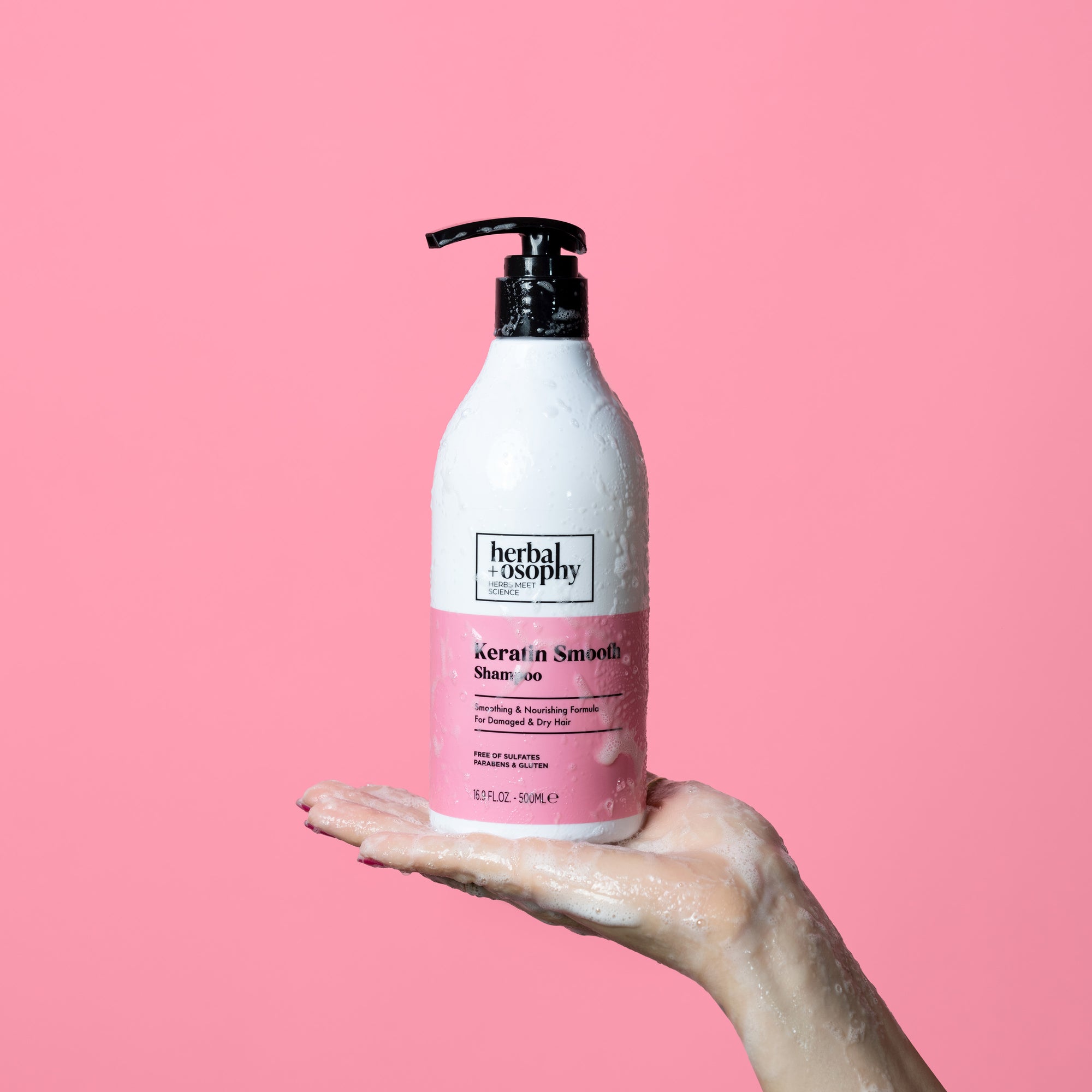 Herbalosophy Keratin Smooth Shampoo bottle held in palm of hand with wet suds on bottle and hand in front of pink background