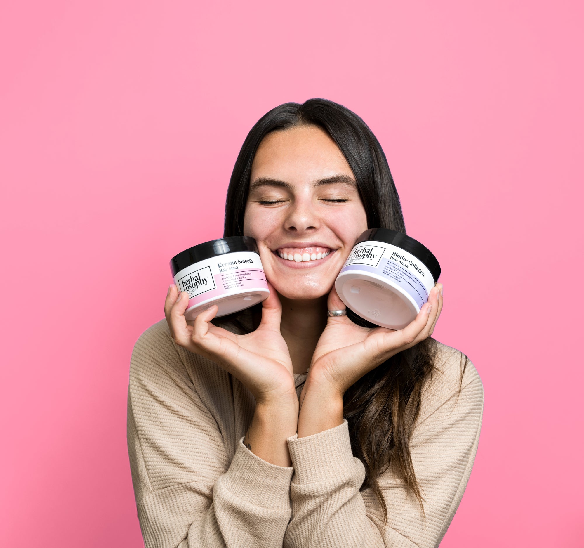woman with long brown hair smiling holding Herbalosophy hair mask jars to her face