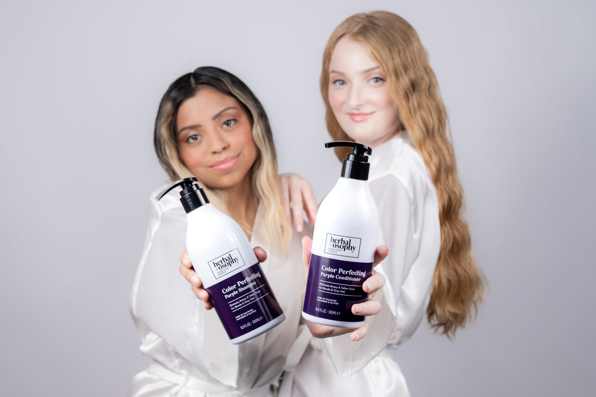 brunette and red haired models in white silk robes holding Herbalosophy Color Perfecting Purple Shampoo and Conditioner bottles out in front of them